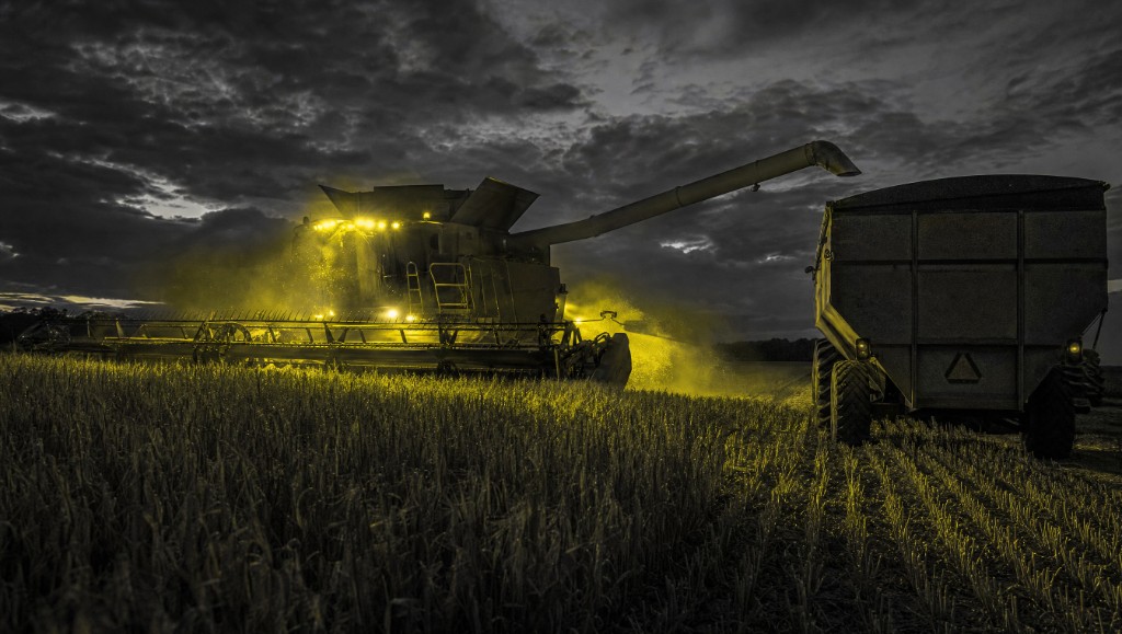 A combine harvester working a grain field under floodlights at dusk, unloading into a waiting cart